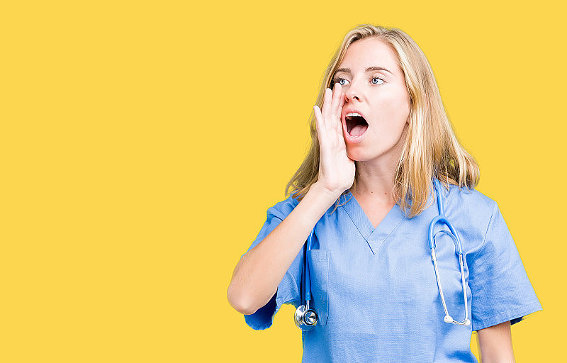 美丽的年轻女医生穿着医疗制服在孤立的背景大声喊叫和尖叫，一边与手在嘴上。通信的概念。