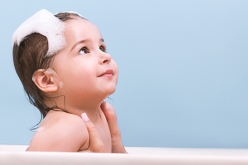 可爱的蹒跚学步的婴儿正在洗澡，玩着泡沫泡泡。浴缸里的小孩抬头看。妈妈带着孩子在蓝色背景的浴室里。婴儿洗澡。卫生和保健。