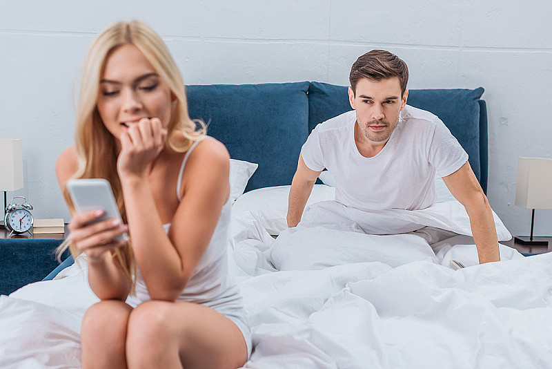 年轻漂亮的女人在用智能手机，嫉妒的男朋友在床上偷看