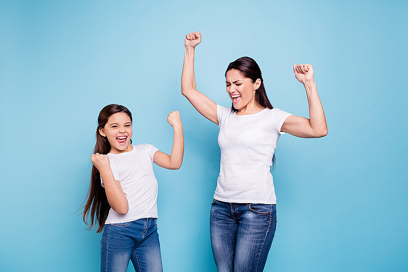 近距离的照片欢呼漂亮的两人棕色头发的妈妈，小的女儿双臂高举在空中高兴地呼喊着家庭游戏的胜利，穿着白色t恤孤立亮蓝色的背景
