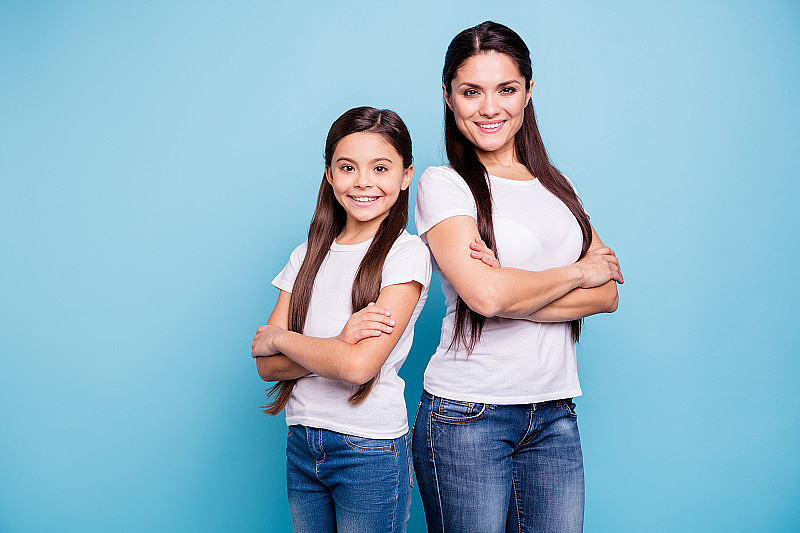 近距离的照片漂亮的两个人棕色头发的妈妈，小的女儿交叉双臂自信地站在背靠背准备赢的家庭游戏穿着t恤孤立的亮蓝色背景