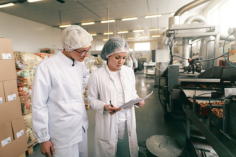 两名身穿白色无菌制服的质量专业人员站在食品厂检查盐棒的质量。