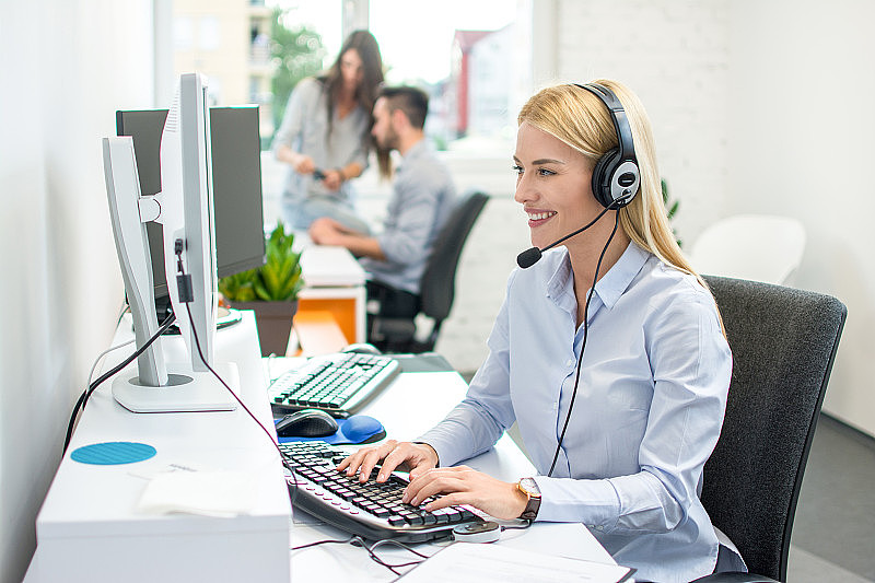 微笑的客户支持女代理人与耳机交谈与客户，同时打字在明亮的办公室和同事在后台。