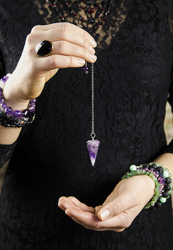 特写的女人穿着黑色哥特式服装，手拿和用紫水晶摆在银链上的算命摆在手里。神秘的巫师读手相的概念。