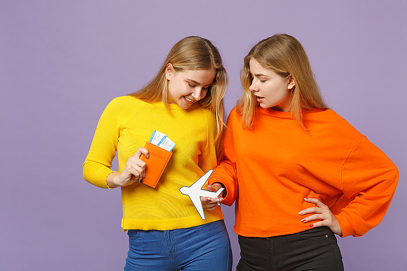 两个漂亮的金发双胞胎姐妹女孩穿着五颜六色的衣服拿着护照、登机牌机票，纸飞机孤立在紫蓝色的背景上。人的家庭生活理念。模拟拷贝空间。