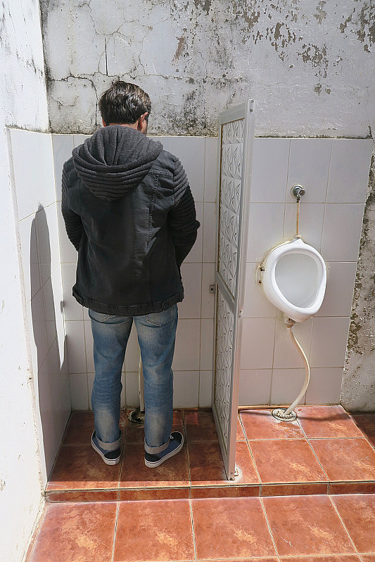 印度男子在户外便池小便，男子在公共厕所小便，印度德里附近的街道，穿着蓝色牛仔牛仔裤，黑色连帽夹克，缺乏厕所便池小便