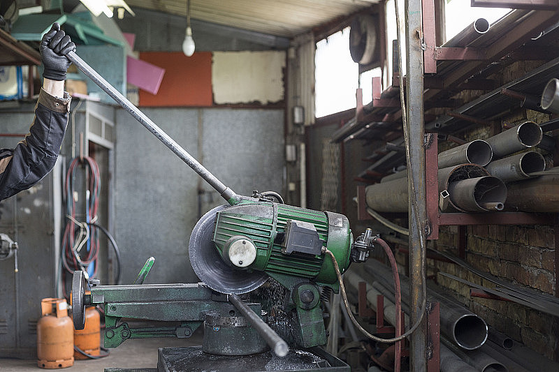 在冶金车间，戴着面罩和手套的工人正在用圆锯切割一根金属管。他穿着防护服。