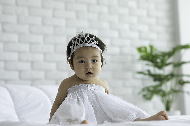 可爱甜美可爱的亚洲宝宝穿着新娘礼服和冠冕坐在白色的床上微笑，在舒适的卧室玩幸福的情绪，健康宝宝的概念