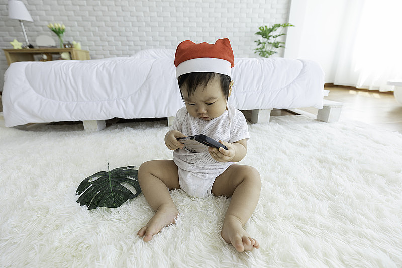 可爱可爱可爱的亚洲宝宝戴着圣诞老人帽服装坐在白色地毯上微笑，在舒适的卧室里玩智能手机或手机与幸福情绪，健康宝宝概念
