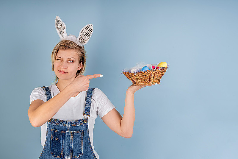 一个长着兔子耳朵的年轻女子指着一个篮子，篮子上的复活节彩蛋被孤立在蓝色的背景上