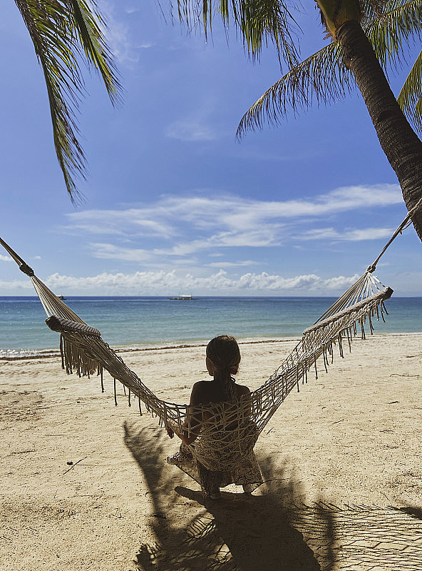 一个没有脸的女孩正坐在白色沙滩上的吊床上。一个女人在后面看着碧蓝的大海和蓝天。一个人正在美丽的度假胜地休息。