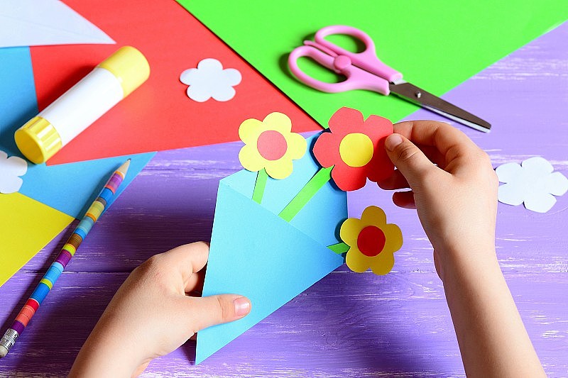 小孩子为母亲节或生日制作纸工艺品。小孩在给妈妈做纸花。简单又漂亮的礼物主意。剪刀，胶水，花模板，铅笔放在木桌上