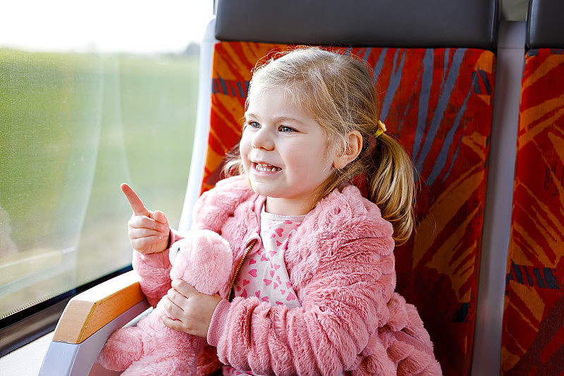 可爱的小女孩坐在火车上，看着窗外，而移动。可爱、快乐、健康的宝宝手里拿着毛绒玩具。笑容满面的孩子要坐火车全家度假