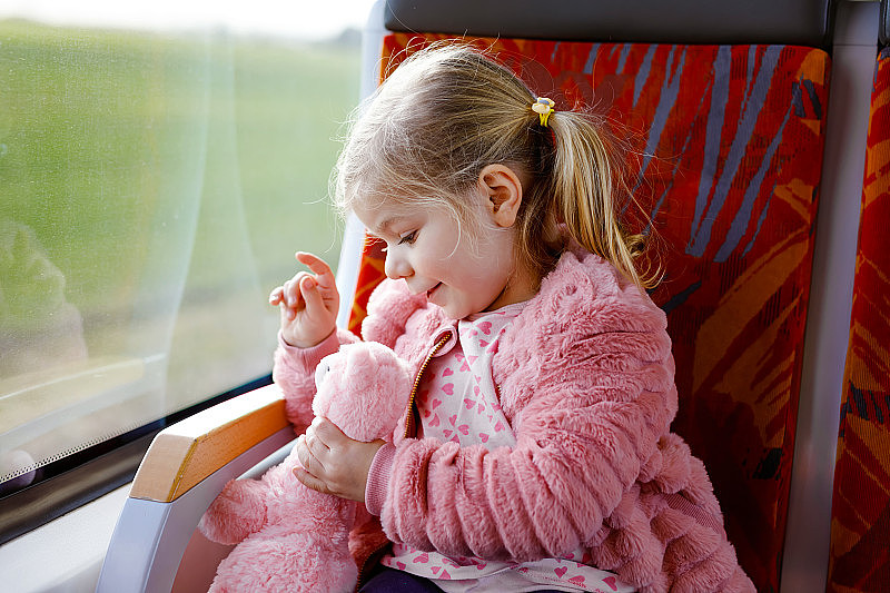 可爱的小女孩坐在火车上，看着窗外，而移动。可爱、快乐、健康的宝宝手里拿着毛绒玩具。笑容满面的孩子要坐火车全家度假