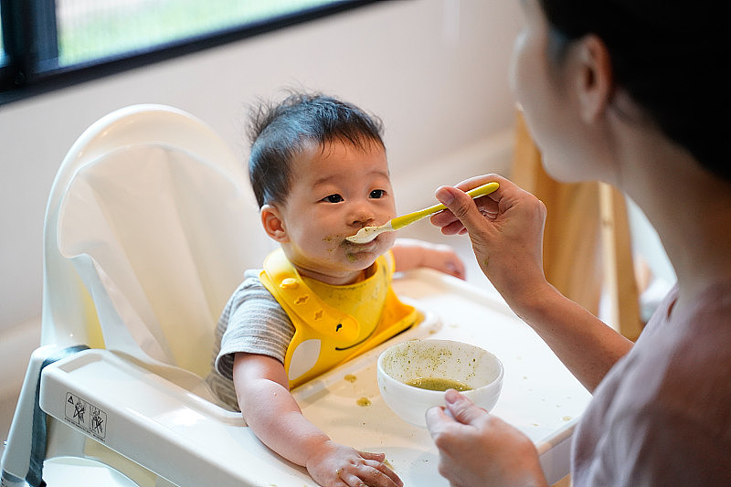 妈妈坐在高椅上喂宝宝，开始给宝宝喂固体食物。小女孩吃蔬菜和儿童健康营养。父母哺育孩子的观念。