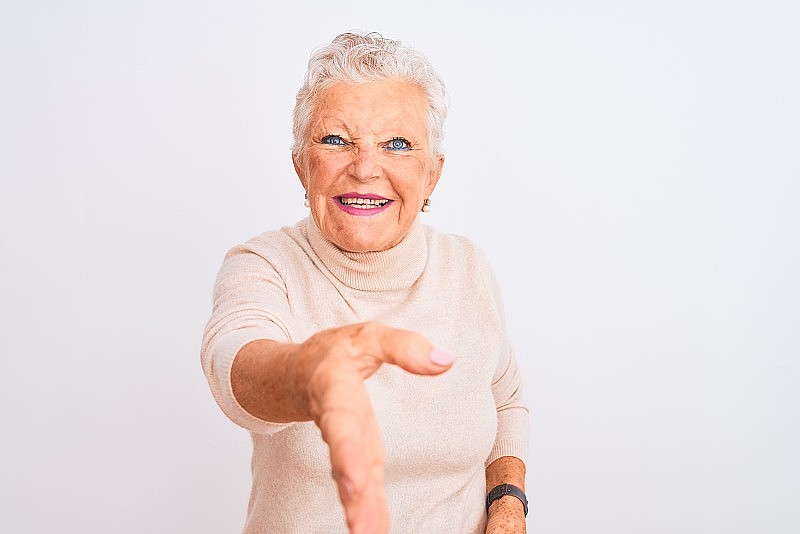 年长的灰色头发的妇女穿着高领毛衣站在孤立的白色背景微笑友好地提供握手作为问候和欢迎。成功的事业。