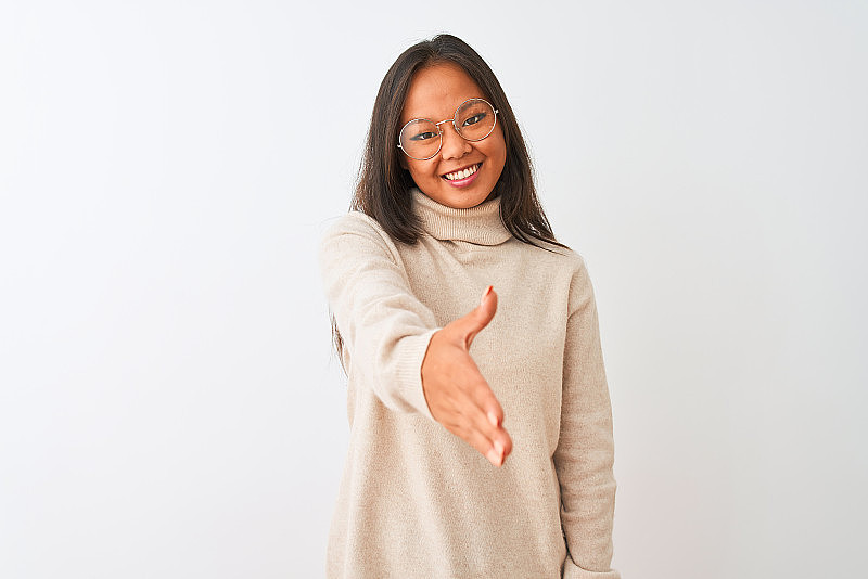 年轻的中国妇女穿着高领毛衣和眼镜在孤立的白色背景微笑友好地提供握手作为问候和欢迎。成功的事业。