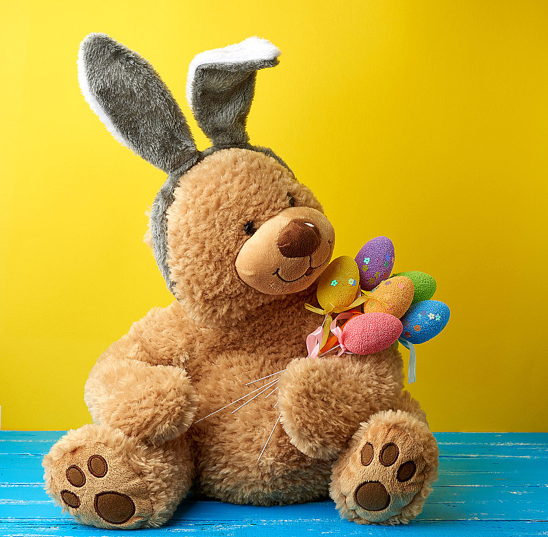 可爱的棕色大泰迪熊拿着五颜六色的复活节彩蛋，头上戴着一个长耳朵的兔子面具