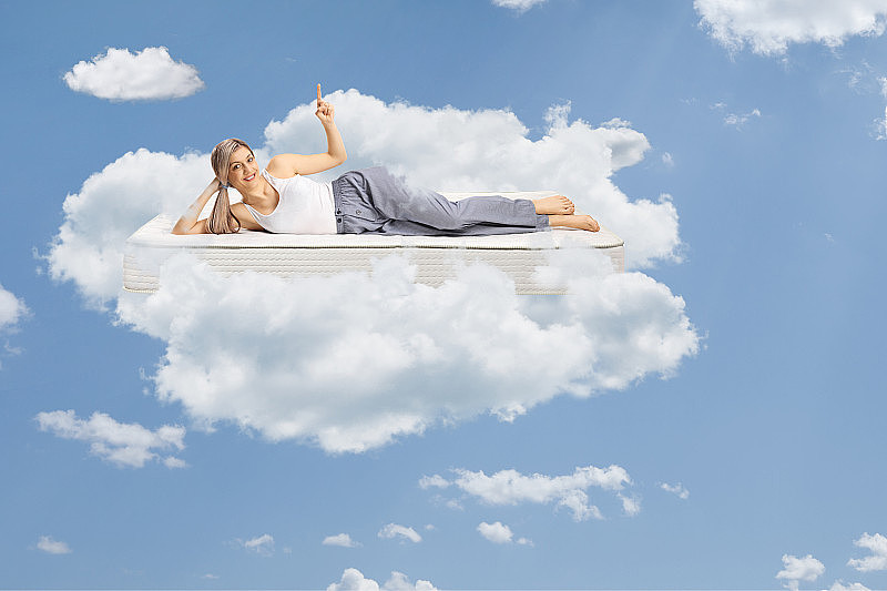 年轻女子躺在一个舒适的床垫上，穿着睡衣，指着天空，漂浮在云上