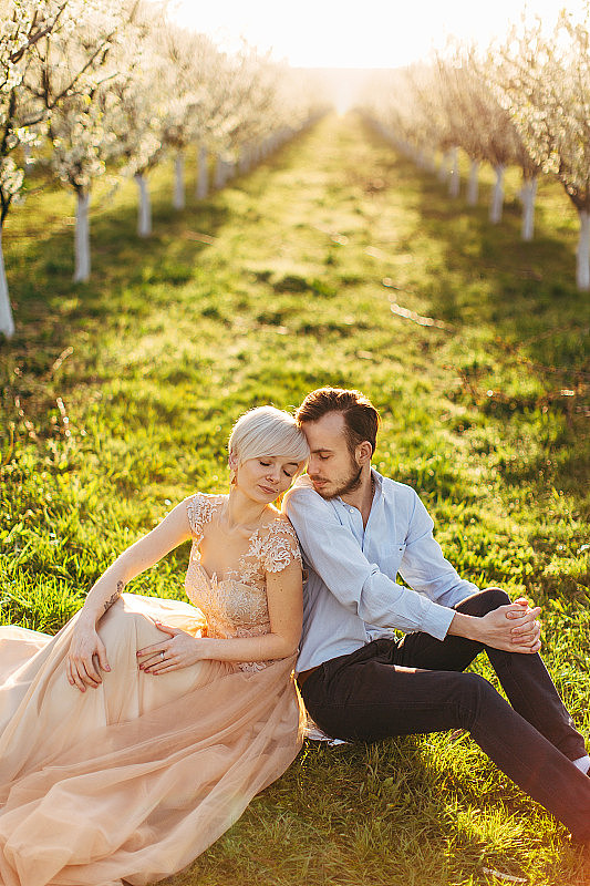年轻可爱的夫妇背靠背坐在绿色的草地上，背景是美丽的日出和开花的树木在花园里。穿着长裙的漂亮女人靠在她那长胡子的英俊男人身上