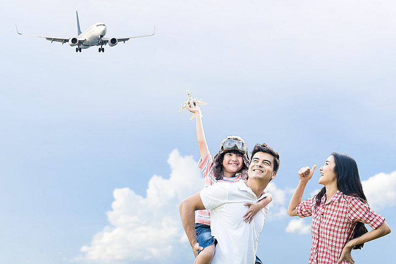 爸爸背着戴着机长帽的男孩，和妈妈一起在空中玩飞机玩具。小孩子的梦想是长大后能成为一名飞行员。家人支持儿子的理想工作或职业。