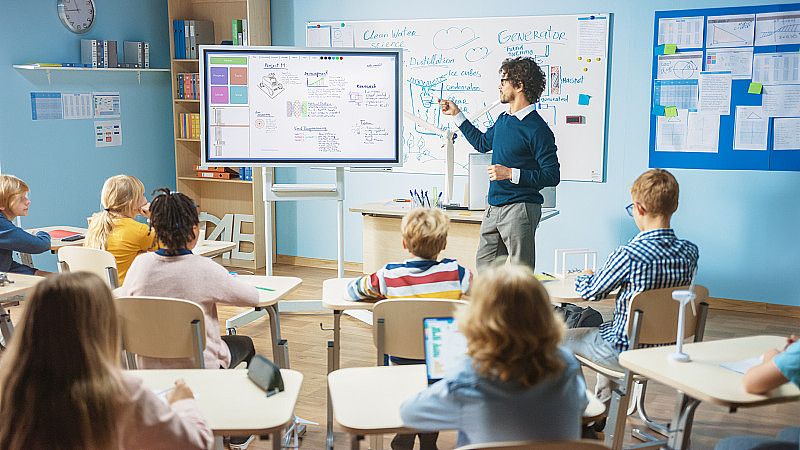 小学科学教师使用交互式数字白板向满教室的孩子展示软件编程如何为机器人工作。科学课，好奇的孩子用心听