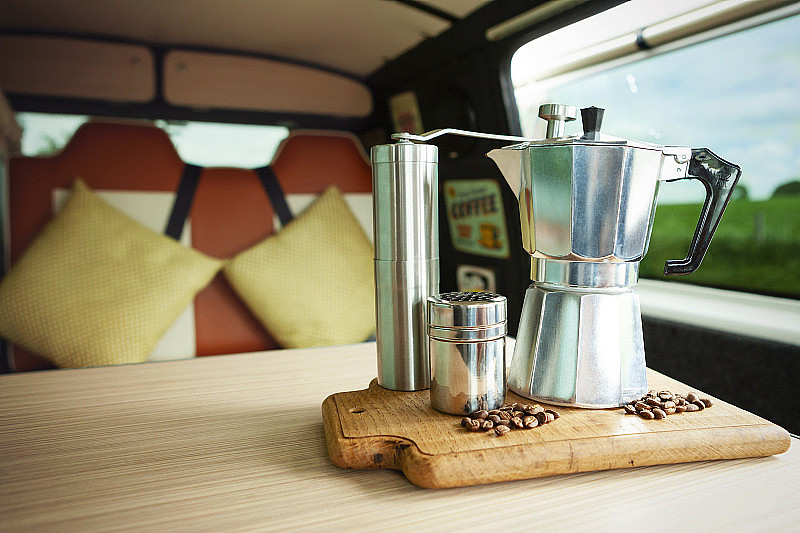 Aqua Bialetti炉顶咖啡机，咖啡研磨机，巧克力摇瓶和咖啡豆，在一辆舒适的露营车里，在一个木制托盘上准备咖啡