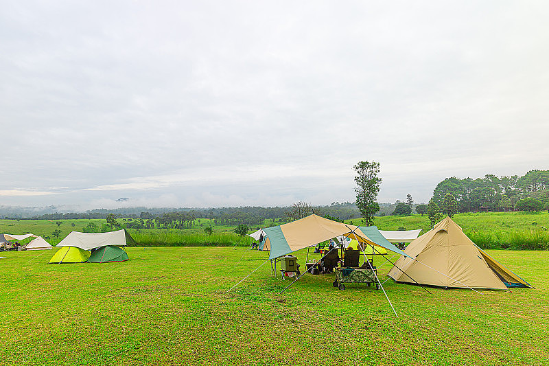 大自然露营帐篷与树在绿色的草地和山在丛林花园与雾在蓝天和露营的人坐在椅子上家庭度假野餐假日放松旅行在温暖的早晨