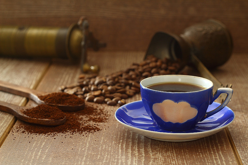 咖啡豆和磨碎的咖啡放在木勺里，棕色的背景。有咖啡壶，蓝色咖啡杯和咖啡研磨机。