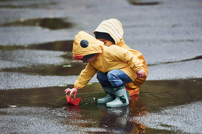 雨后，两个穿着黄色防水斗篷和靴子的孩子一起在户外玩手工纸船