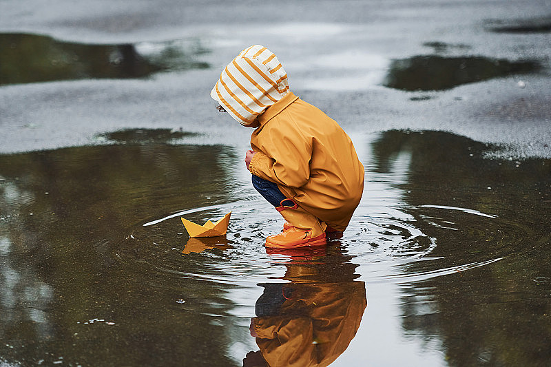 雨后，穿着黄色防水斗篷和靴子的孩子在户外玩纸手工船玩具