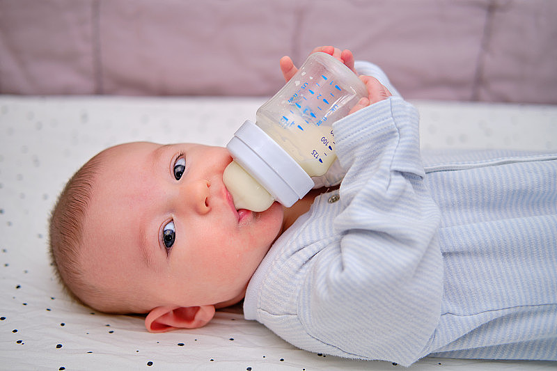 快乐的宝宝手里拿着一瓶牛奶，独自微笑着。三个月大的孩子自己躺在床上