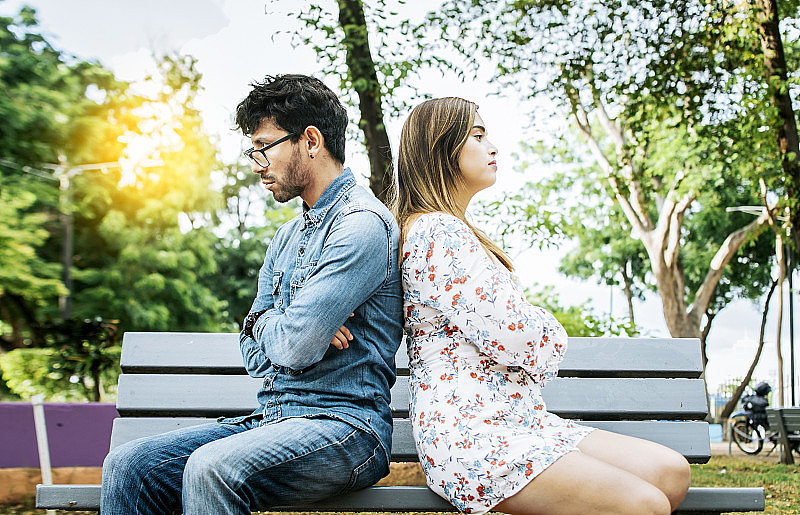 心烦意乱的年轻夫妇背靠背坐在公园里，不开心的夫妇背靠背坐在长椅上。夫妻问题和危机的概念。一对年轻夫妇背靠背坐在长椅上争吵