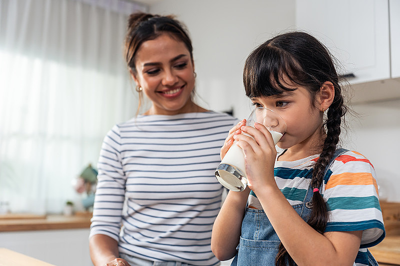 白人小孩拿着一杯牛奶和妈妈一起喝。迷人的妈妈教导和支持年轻的女孩照顾自己的身体，醒来后在家里喝牛奶进行保健。