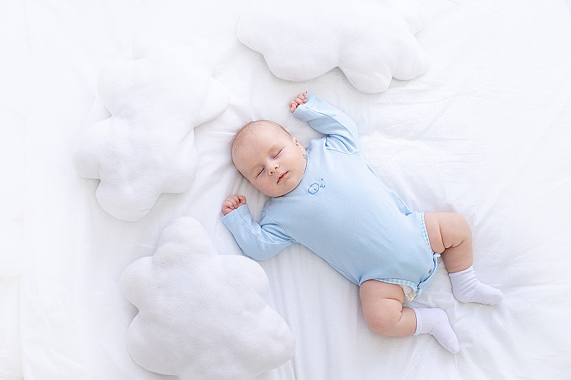 男婴穿着蓝色睡衣躺在床上，双手高举在云雾的枕头间，健康地睡着新生儿