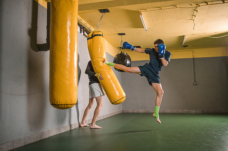 一名年轻的男子自由搏击运动员在健身房通过踢他的朋友拿着的沙袋进行训练