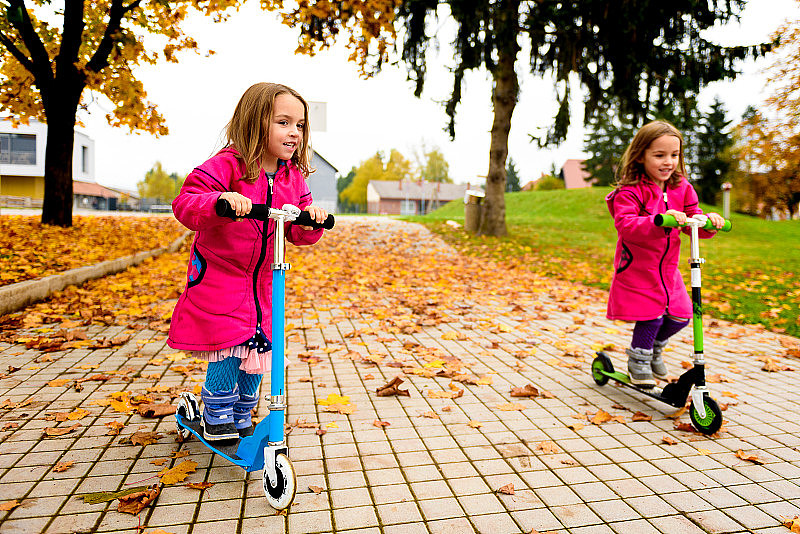 一对穿着粉红色外套的双胞胎女孩骑着踏板车在枫叶上。