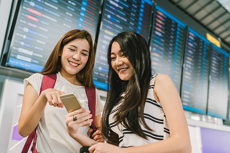 两个亚洲女孩在机场的航班信息板上一起使用智能手机。在线登记，时间表申请，或假日旅游概念