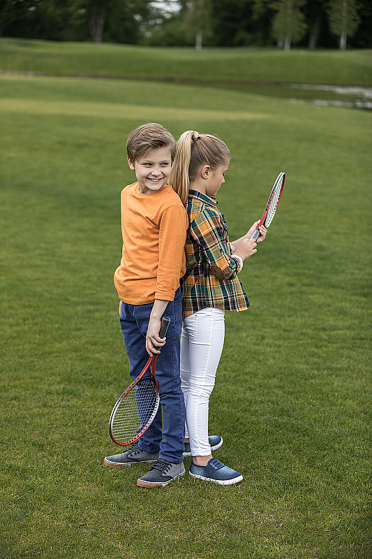 可爱的男孩和女孩拿着羽毛球拍背靠背站在绿色的草地上