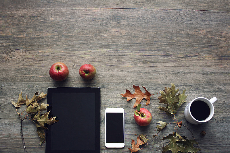 秋天的静物，红色的苹果，移动设备，黑色的咖啡杯和秋天的树叶在乡村的木制背景