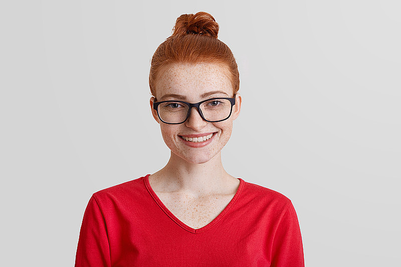 工作室拍摄的红头发年轻微笑的女性戴着方形眼镜和红色毛衣，在工作中升职后心情很好，因工作勤奋获得奖励，取得了巨大的成功