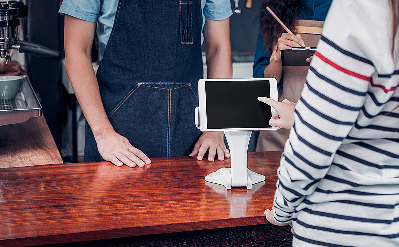 顾客在咖啡厅柜台吧台用平板电脑屏幕自助点餐，卖家咖啡店接受手机付款。数字生活方式的概念。设计展示的空白。剪切路径。