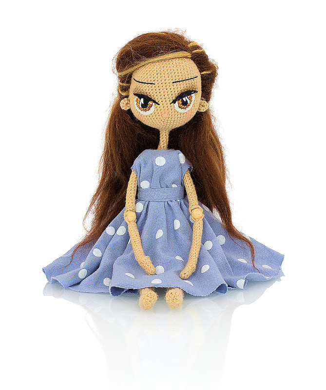 可爱的手工钩针娃娃与斑点蓝色连衣裙孤立在白色背景与阴影反射。好玩的钩针棕色头发娃娃坐在白色的衬里。针织的洋娃娃。