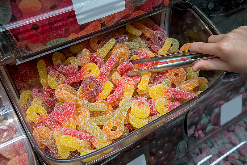 在糖果店，小孩用钳子在塑料容器中挑选泡沫和酸味软糖。
