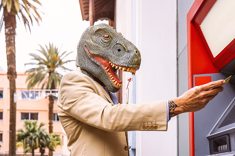 戴着霸王龙恐龙面具的老人用借记卡从银行取款机取款-半人半兽的超现实形象- ATM广告的荒谬和疯狂概念