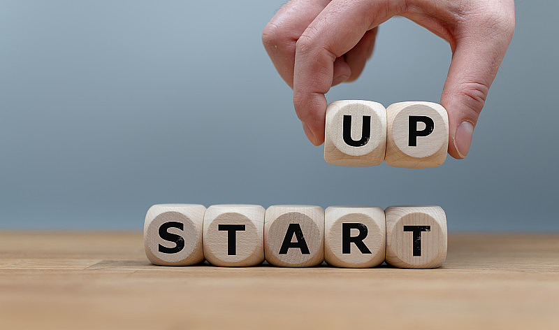 立方体组成单词“START UP”，同时手指在空中举起字母“UP”。立方体摆放在灰色背景前面的一张木桌上。