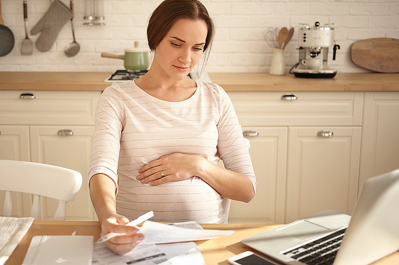 室内拍摄的忙碌认真的年轻孕妇保持一只手在肚子上，拿着一张纸，用笔记本电脑在线支付账单，已经集中注意力，坐在厨房里