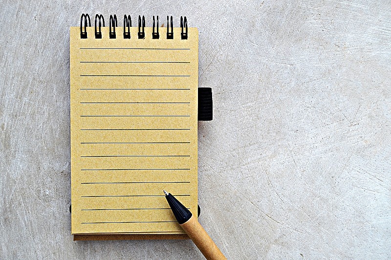 一个单行空白的黄色米色页，一个螺旋装订的垂直记事本和棕色和黑色的钢笔放在上面，上面是一个木制的米色陈旧的老式水平背景。