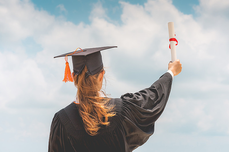 毕业女生戴着学士帽和学士服，手拿证书，蓝天背景，背景，暑期教育成功理念