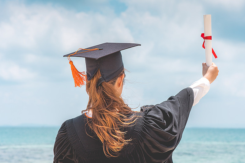 毕业女生戴着学士帽和学士服，手拿证书，蓝天背景，背景，暑期教育成功理念
